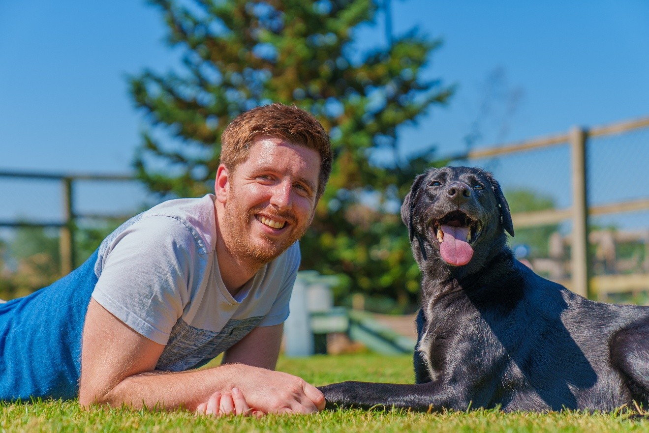 Smiling dog owner lying on grass beside a happy black dog outdoors, showing a calm and relaxed bond between pet and owner.