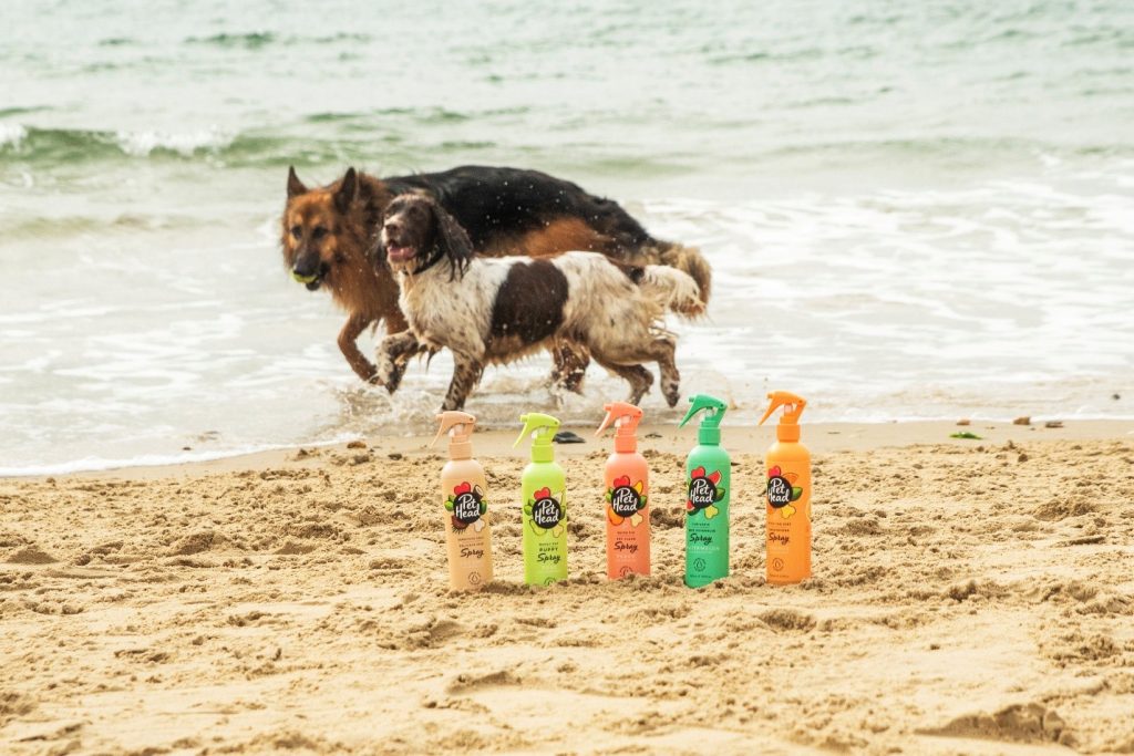 Lineup of Pet Head dog sprays and grooming products on a sandy beach with dogs playing in the background.